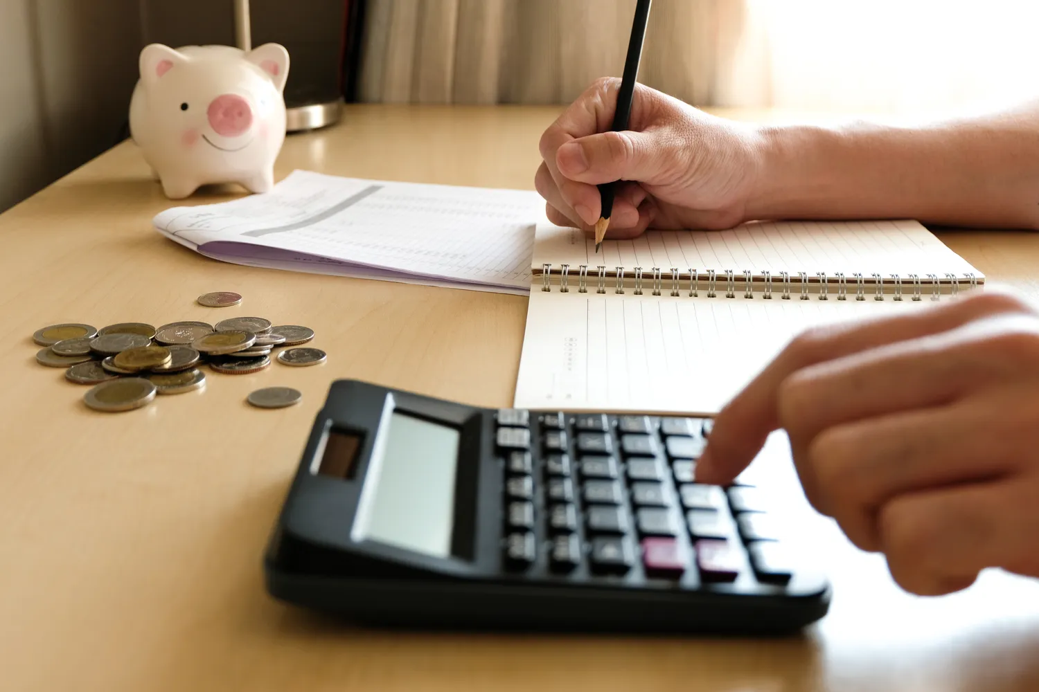 Man writing on paper and working with a calculator near a piggy bank to demonstrate the concept of cost